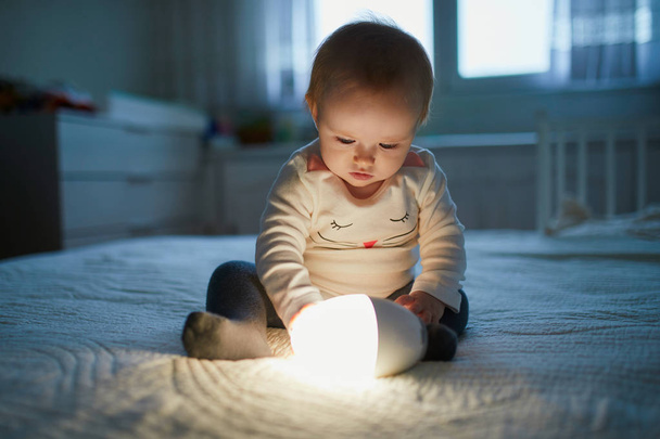 Adorable baby girl playing with bedside lamp in nursery - Zdjęcie, obraz