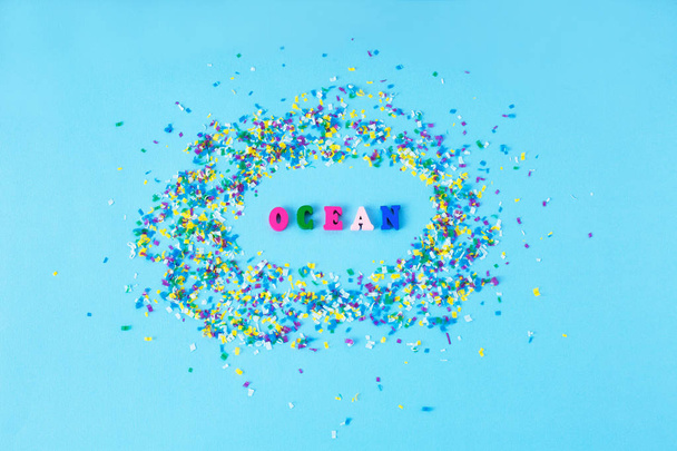Ξύλινα γράμματα με λέξη ωκεανού γύρω από μικρά πλαστικά μόρια σε μπλε φόντο. Microplastic πρόβλημα. Επικίνδυνα πρόσθετα. Τοξικές ουσίες. - Φωτογραφία, εικόνα