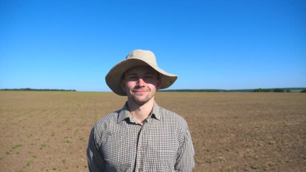 Porträt eines glücklichen jungen Bauern mit Hut, der in die Kamera blickt und vor dem Hintergrund von Feld und blauem Himmel lächelt. Nahaufnahme eines gutaussehenden Mannes im Hemd, der an einem sonnigen Sommertag auf der Wiese steht - Filmmaterial, Video