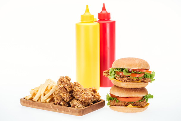 хрусткі курячі нагетси, гамбургери та картопля фрі біля пляшок з кетчупом та гірчицею ізольовані на білому
 - Фото, зображення
