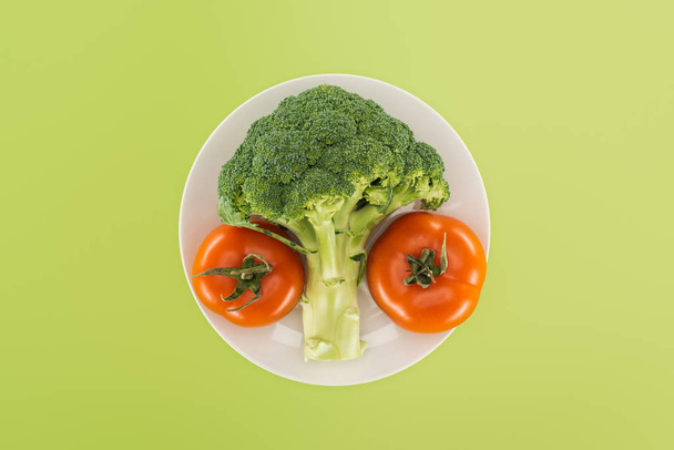 top view of organic ripe broccoli near red tomatoes on white plate isolated on green - Photo, Image