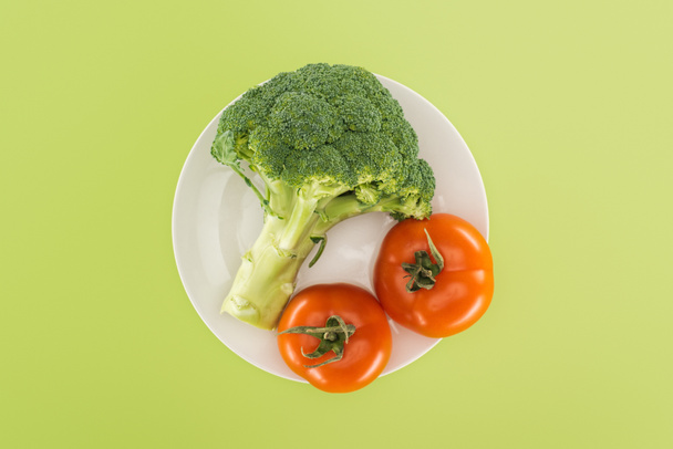 top view of organic broccoli near red tomatoes on white plate isolated on green - Photo, Image