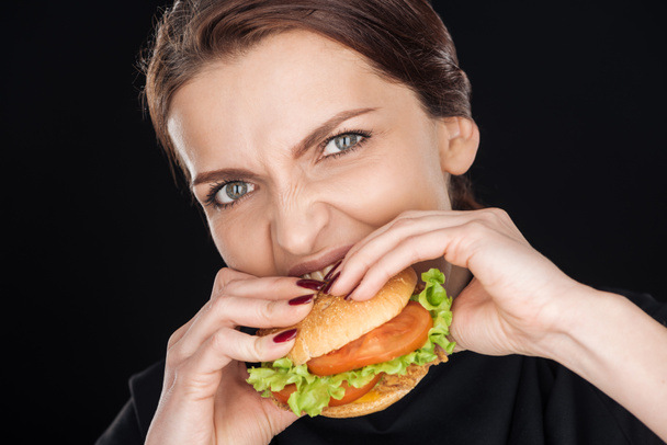 Irritierte Frau isst leckeren Chicken Burger, während sie isoliert auf schwarz in die Kamera schaut - Foto, Bild