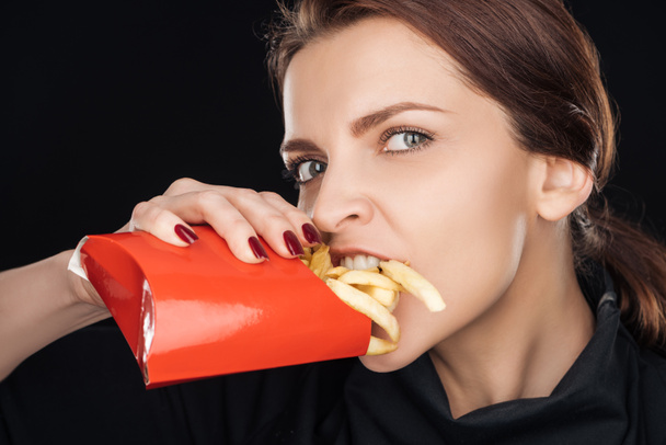 atraktivní žena jíst slané francouzské hranolky při pohledu na fotoaparát izolované na černém pozadí - Fotografie, Obrázek