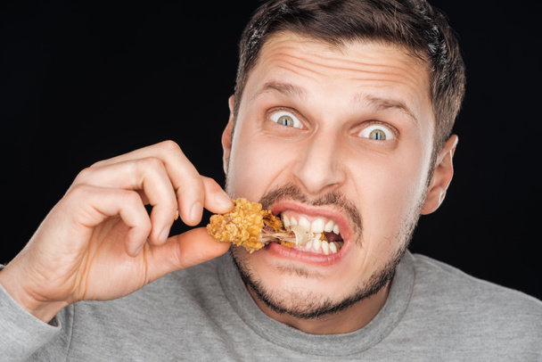 emotional man eating crispy chicken nugget while looking at camera isolated on black - Foto, Imagem
