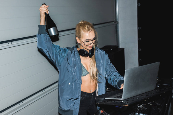 cheerful dj woman holding bottle in hand and looking at laptop - Фото, изображение