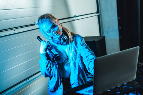 atractiva mujer dj en gafas sosteniendo botella mientras está de pie cerca del ordenador portátil
 - Foto, imagen