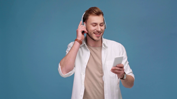 Mann mit Kopfhörer hört Musik auf Smartphone und lächelt isoliert auf blauem Grund - Filmmaterial, Video