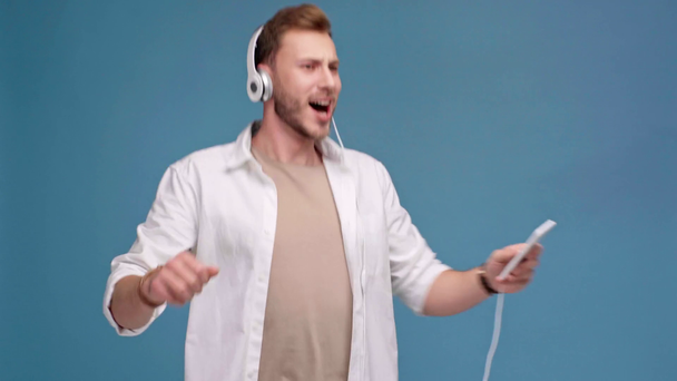 handsome happy man in headphones listening music on smartphone and dancing isolated on blue - Video