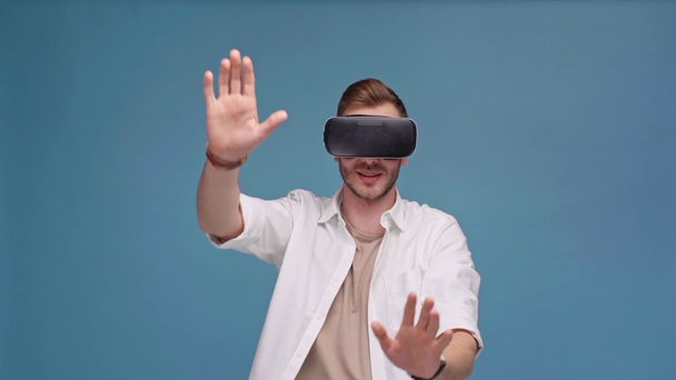 człowiek w zestaw wirtualnej rzeczywistości gestykulacji z rąk i uśmiecha się na białym tle na niebiesko - Materiał filmowy, wideo
