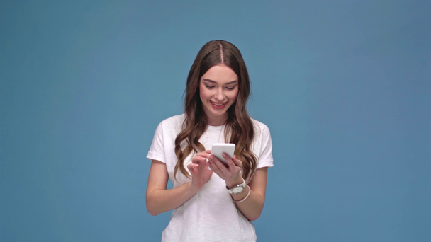 beautiful girl using smartphone and smiling isolated on blue with copy space - Video, Çekim