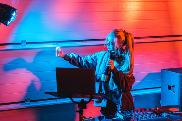 atraktivní blondýna dj žena držící láhev a pořizování selfie v nočním klubu - Fotografie, Obrázek