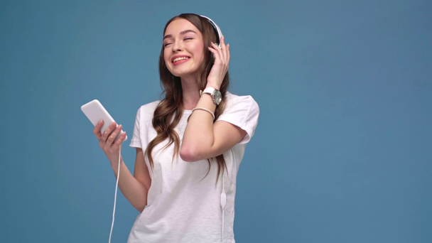 krásná dívka ve sluchátkách poslouchat hudbu na smartphone, tančí a usmívá se izolované na modré - Záběry, video