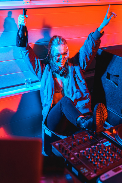 vrolijke dj vrouw in hoofdtelefoons houden fles en gebaren tijdens de vergadering in nachtclub - Foto, afbeelding