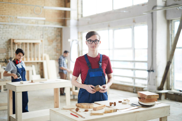 Fiatal Ács szemüveg viselése a tartalom és a kék egységes állandó asztalnál és a gazdaság fából készült darab fa modern gyári munka közben - Fotó, kép