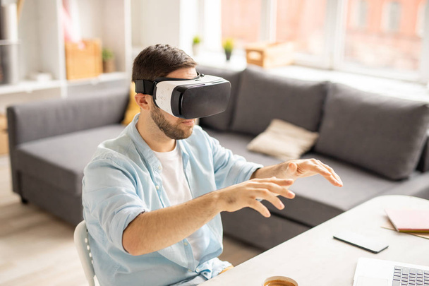 Contemporary businessman in vr goggle sitting by desk in front of virtual display and looking at stuff on it - Photo, Image