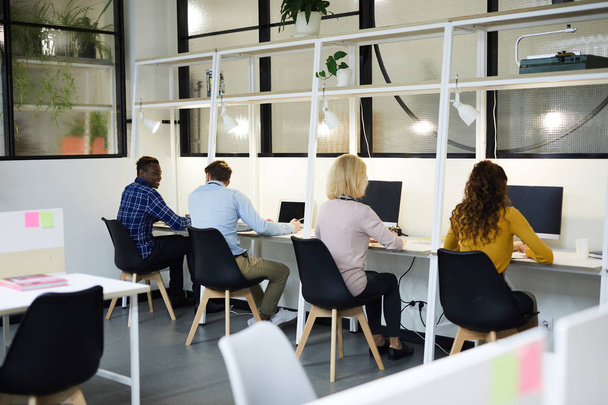 Vista trasera de las personas de negocios multiétnicos en ropa casual sentados en escritorios con computadoras y trabajando con información, personas en fila
 - Foto, imagen