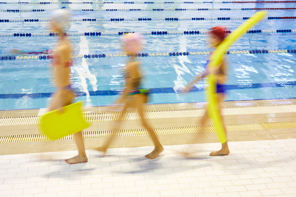 水泳レッスンでプールサイドを歩く水泳麺やボードを持つ子供たちのグループのぼやけた動き - 写真・画像