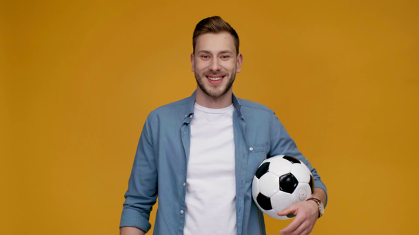 handsome man spinning soccer ball on finger, smiling, looking at camera and showing thumb up sign isolated on yellow - Felvétel, videó