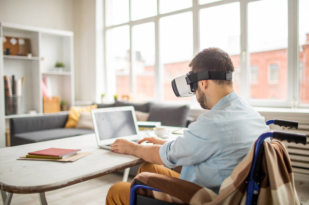 Empresário com óculos VR sentado em cadeira de rodas na frente do laptop e rede enquanto olha para a exibição virtual
 - Foto, Imagem