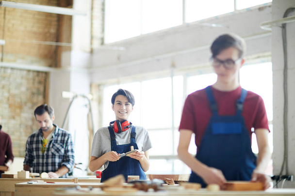 Cheerful attractive young lady with ear protectors on neck standing at desk and measuring wooden detail while working in carpentry shop - Photo, Image