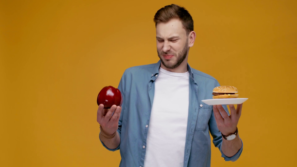 knappe man bedrijf voedsel in handen, aroma van Hamburger ruiken, kijken camera en glimlachend geïsoleerd op geel - Video