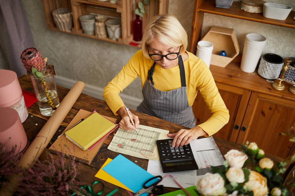 Retrato de ángulo alto de la mujer de negocios contando las finanzas usando calculadora en pequeña tienda, espacio de copia
 - Foto, Imagen