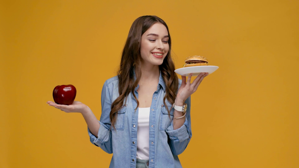 beautiful girl holding food in hands, biting lip, showing burger at camera and smiling isolated on yellow - Footage, Video