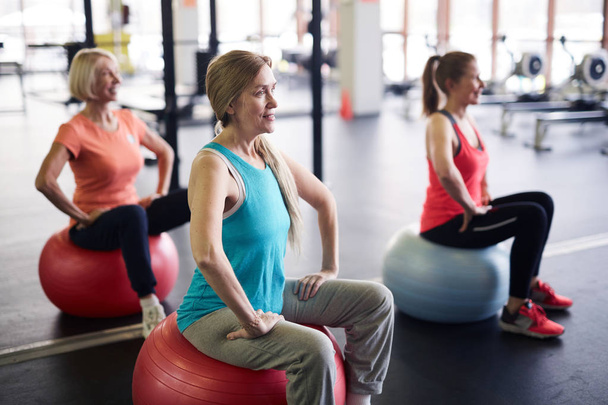 Group of mature active females in sportswear sitting on fitballs and listening to their instructor before training - Foto, imagen