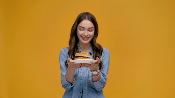 krásná šťastná dívka kousání rtu, při pohledu na fotoaparát a ukazující deska s burger izolované na žluté - Záběry, video