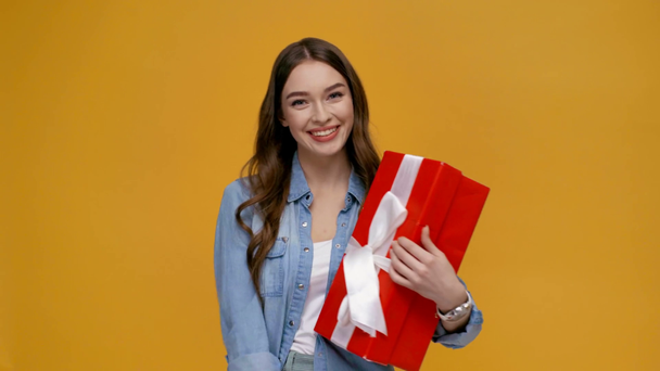 beautiful girl pointing with finger at gift box, smiling, looking at camera and cheering with raised fist isolated on yellow - Footage, Video
