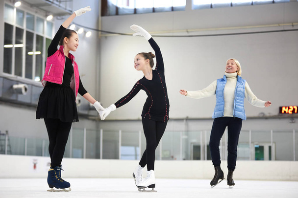 Retrato de longitud completa de dos hermosas chicas patinaje artístico en pista de hielo con entrenador femenino observándolos en el fondo, espacio de copia
 - Foto, Imagen