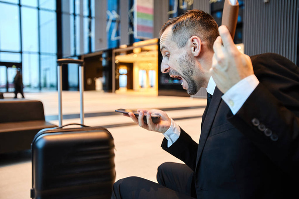 Öfkeli işadamı havaalanı salonunda otururken akıllı telefondan birine bağırıyor - Fotoğraf, Görsel