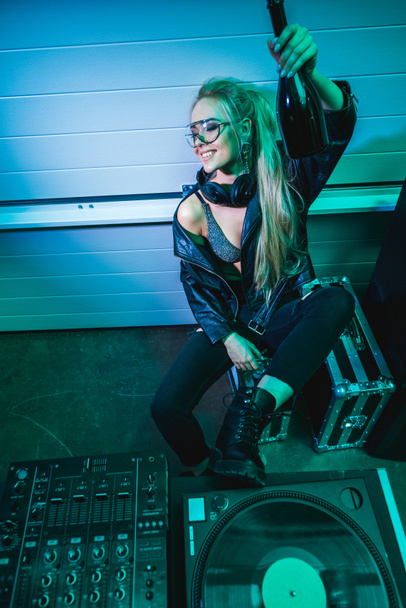 fröhliches DJ-Mädchen in Gläsern, Flasche mit Wein in der Hand und lächelnd im Nachtclub  - Foto, Bild