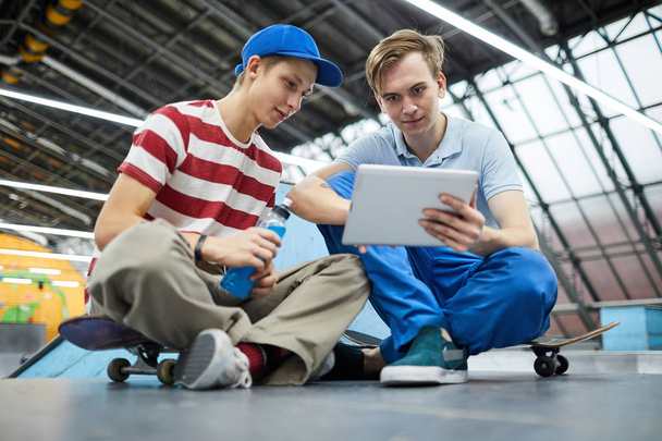 Deux garçons amicaux en tenue décontractée assis sur le sol après la formation de parkour et regarder la vidéo en ligne dans la tablette
 - Photo, image