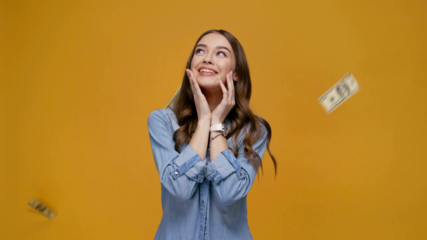 happy girl doing please gesture and looking at falling dollar banknotes isolated on yellow - Footage, Video