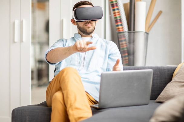 Young businessman with vr headset pointing at virtual display or pressing button while sitting in front of laptop - 写真・画像