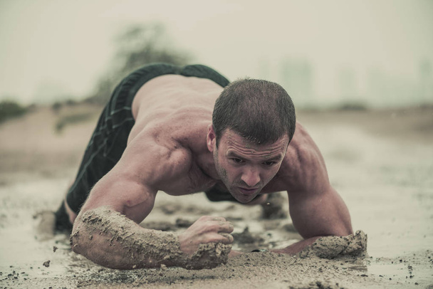 Closeup of strong, muscular, athletic man crawling in a wet, dirty muddy puddle in the rain in an extreme competitive sport  - Foto, afbeelding