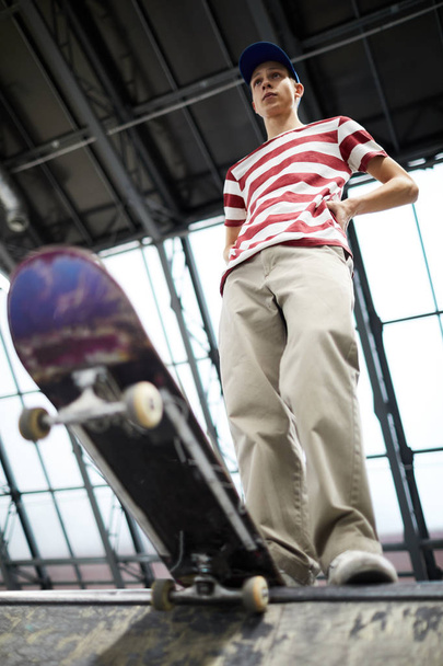 Teenage guy in striped tee and beige pants standing on edge of parkour area before descending on skateboard - Fotó, kép
