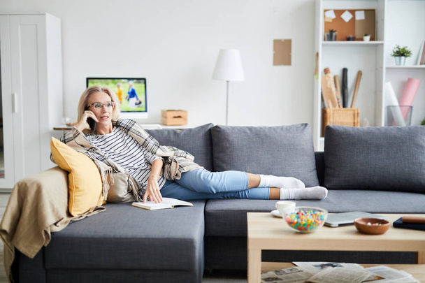 Porträt einer zeitgenössischen Frau, die telefoniert, während sie es sich zu Hause auf einer bequemen Couch bequem macht, Kopierraum - Foto, Bild
