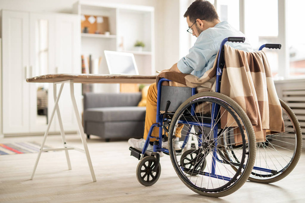 Biznesmen dorywczo na wózku inwalidzkim, organizowanie dnia pracy, siedząc przy biurku w salonie w domu - Zdjęcie, obraz