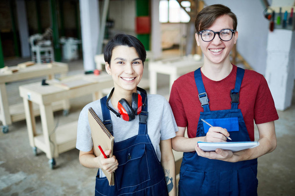 Cheerful satisfied young students in workwear standing in modern carpentry classroom and laughing while looking at camera and writing down information in sketchpad - Фото, зображення