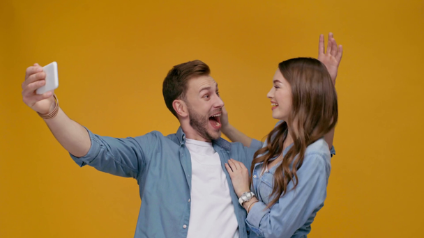 舌を突き出して、黄色の分離された平和の兆しを見せているスマート フォンで selfie を取って美しい幸せなカップル - 映像、動画