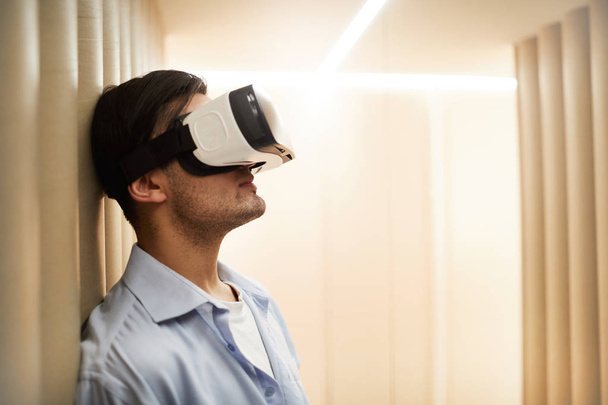 Współczesny młody człowiek z Gogle VR pochylony przez ścianę podczas podróży w wirtualnym świecie - Zdjęcie, obraz