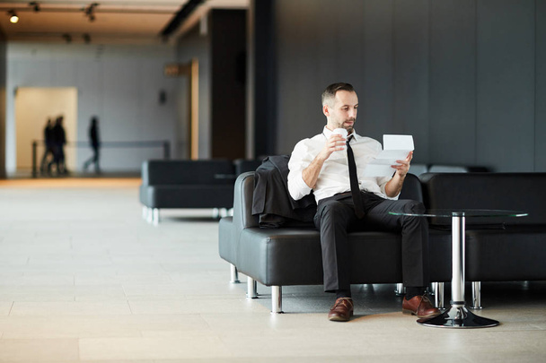 Nuori rauhallinen liikemies tai matkustaja istuu sohvalla lentokentän loungessa, ottaa juoda ja lukea asiakirjan
 - Valokuva, kuva