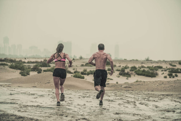 Arkadan görünüş güçlü, kaslı erkek ve kadın sporcuların engebeli bir çöl arka plan aşınmış dokulu bir doğa sporları yarış ile aşağı koşma çamur kaplı bitirmek - Fotoğraf, Görsel