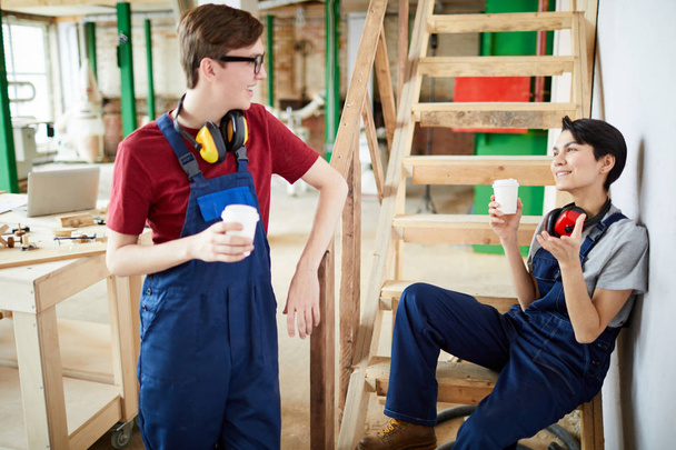 Pozitivní mladých tesařské práce pracovníků v uniformě nosit chrániče sluchu na krku pití kávy a smáli se při chatování během přestávky v tesařství - Fotografie, Obrázek