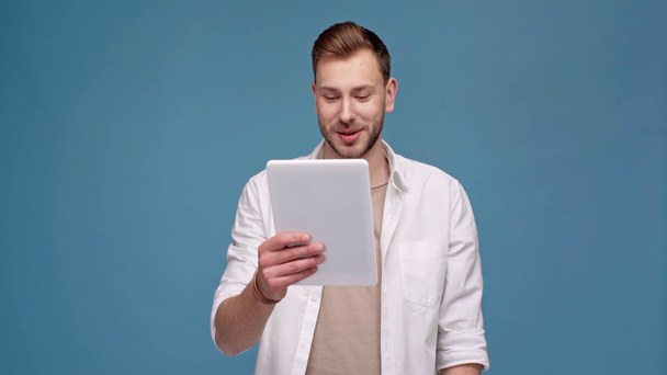 handsome man waving with hand, talking and smiling while using digital tablet during video call isolated on blue - Video, Çekim