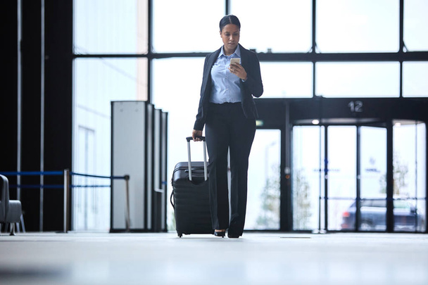 Занятая молодая женщина в формальной одежде, прокручивающая в смартфоне, двигаясь по аэропорту с чемоданом
 - Фото, изображение