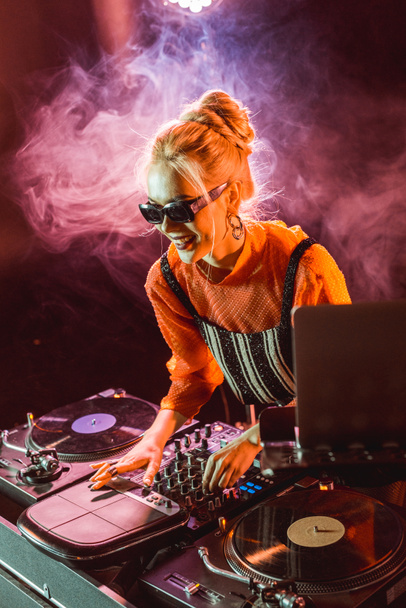 cheerful dj girl in sunglasses touching dj equipment in nightclub with smoke  - Photo, Image